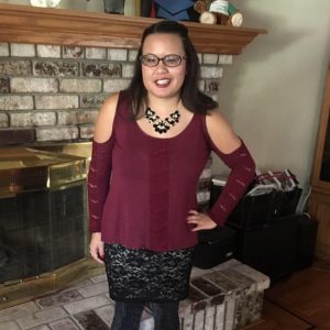 Photo of a light-skinned young asian woman standing and smiling near a fireplace. She is wearing glasses, a maroon blouse and a black skirt.