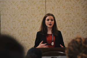 Photo of Kaitlyn (Katy) Brennan, wearing a black blazer and red shirt, speaking to a room.