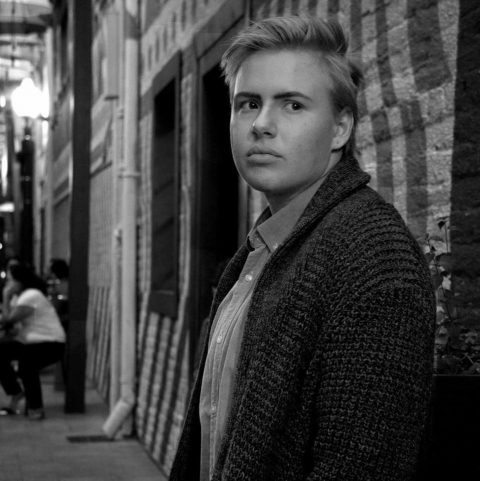 Black and white photo. Young, Caucasian male standing against a wall