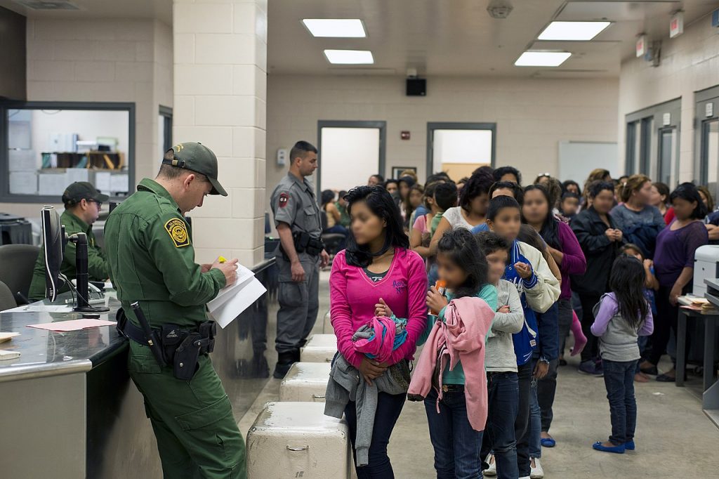 Room full of brown-skinned youth without parents or guardians not smiling in line in a detention center. There are 3 white male guards. 